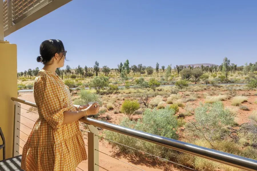 Kvinne på balkongen med utsikt over ørkenbusker og Uluru i det fjerne.