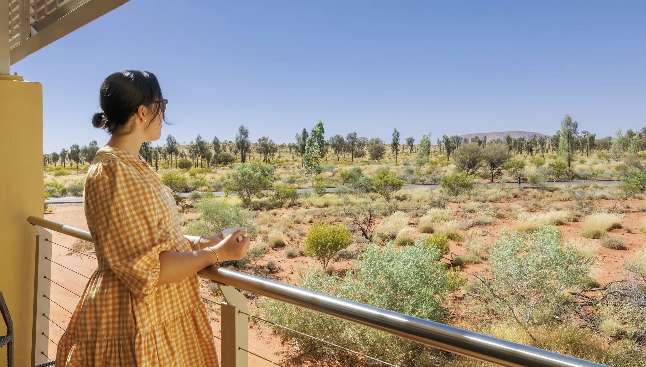 Kvinne på balkongen med utsikt over ørkenbusker og Uluru i det fjerne.