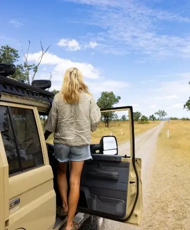 A person is standing inside an open vehicle, looking out as they embark on a journey through a dry, grassy landscape under a clear blue sky.