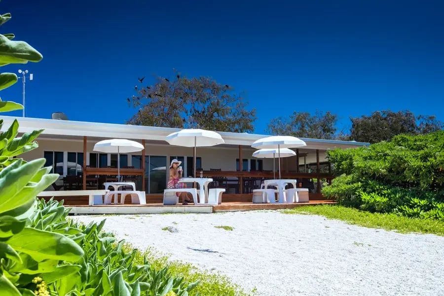 Restaurantbygning med parasoller og uteservering bak hvit korallstrand.