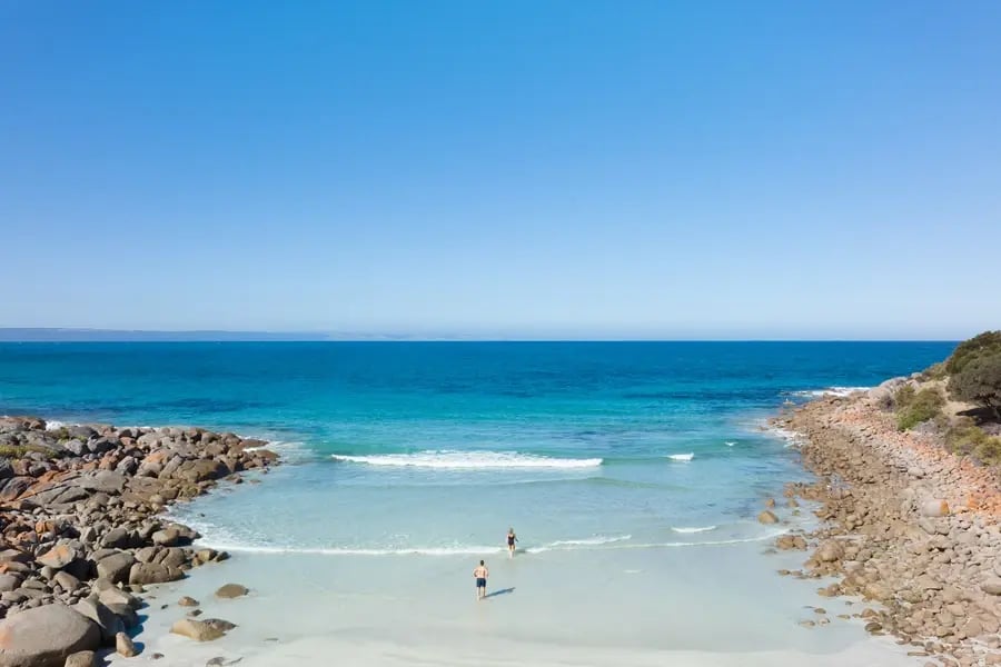 Gjester som bader ved en skjermet strand med klart blått hav og steiner på sidene.