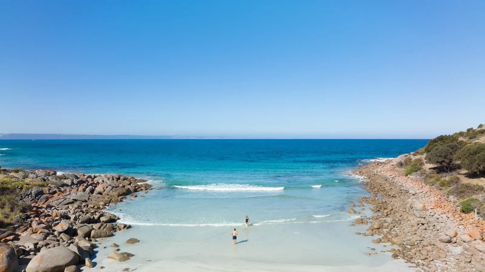 Gjester som bader ved en skjermet strand med klart blått hav og steiner på sidene.