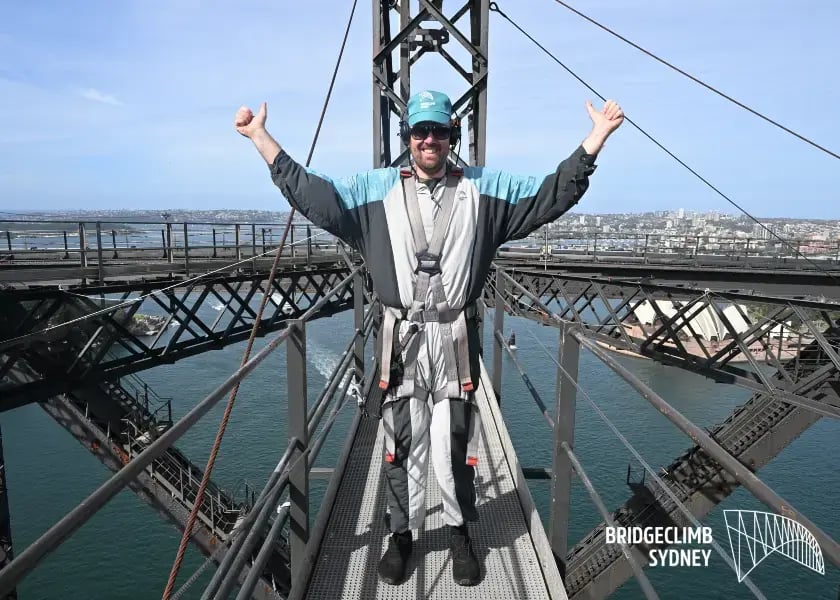 person standing on a steel bridge with their arms raised in a triumphant gesture. They are wearing a helmet, a harness, and climbing gear, suggesting they have just completed a climb. The bridge is named 'BRIDGECLIMB SYDNEY' as indicated by the text on the image
