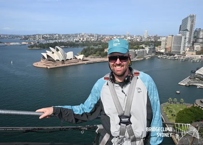 an individual, wearing a blue jacket and a grey backpack, standing with arms outstretched, overlooking the view of the Sydney Opera House and the Sydney Harbour Bridge from a high vantage point. The person appears to be on a bridge or elevated platform, enjoying a clear day with blue skies.