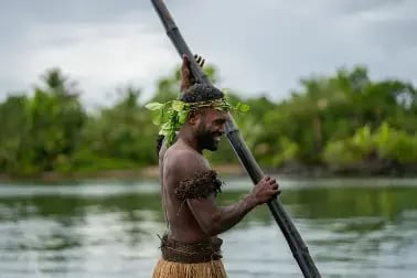 En fijiansk mann står med en stor stokk - kledd i tradisjonelle klær - bastskjørt, plantekrans på hodet og rundt biceps.