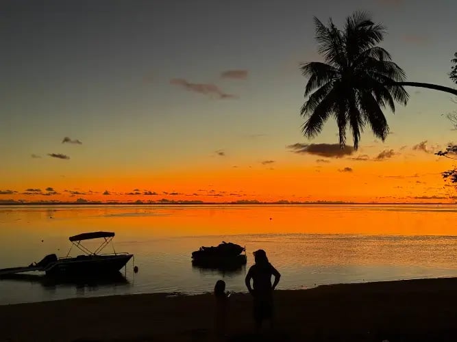 Solnedgang på stranden, med palmer og båter, og personer og en blodrødsol.