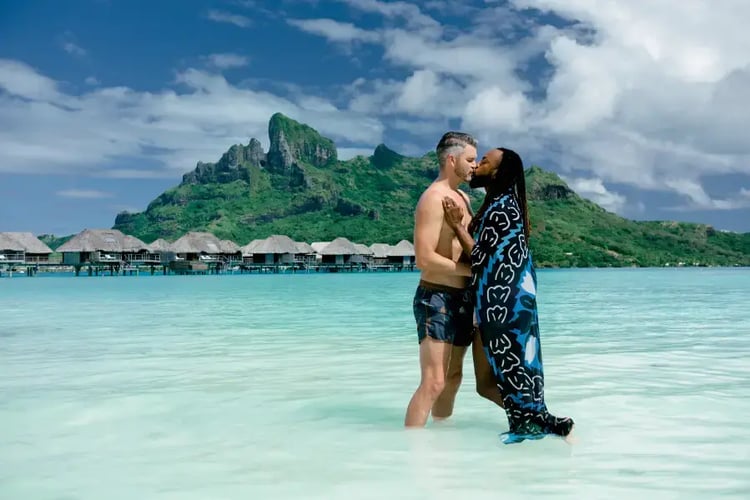 The image shows a couple embracing in a shallow, turquoise body of water with a picturesque mountainous landscape in the background.