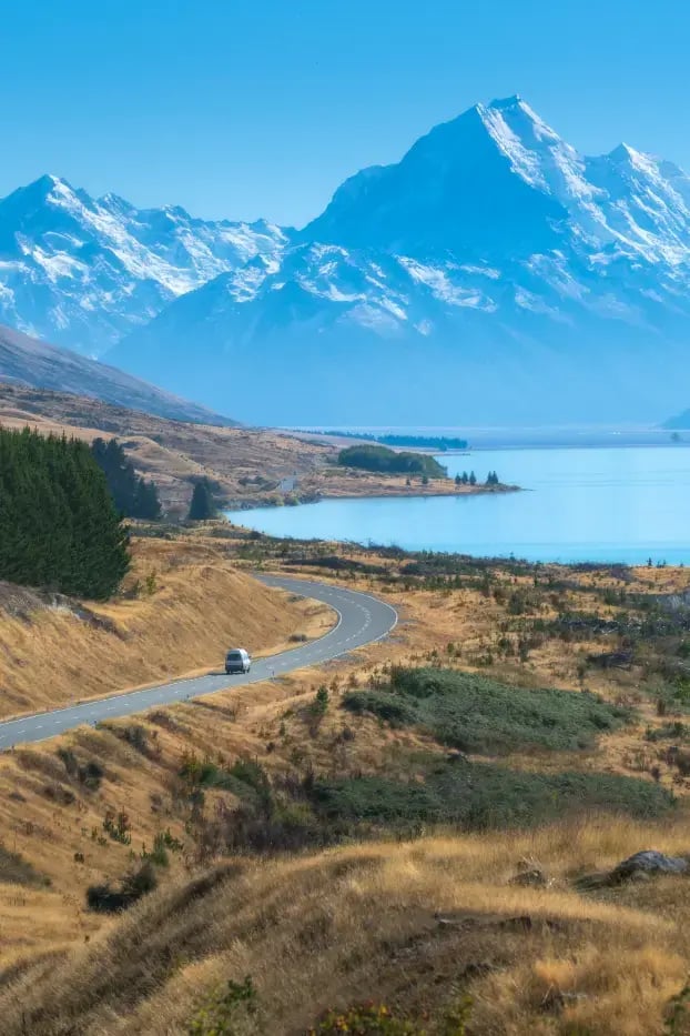 En vei som svinger seg gjennom landskapet,  med innsjø og fjell i bakgrunn. En vil kjører på veien.