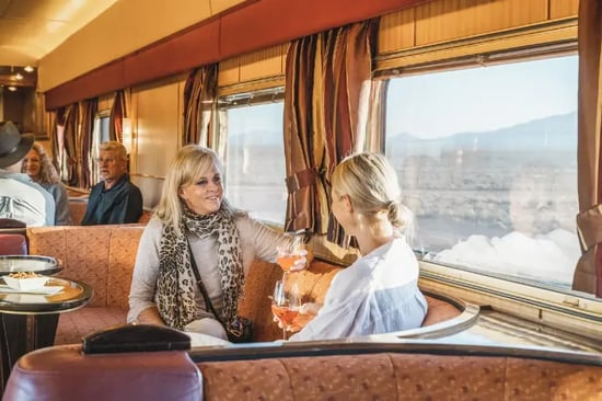 a cozy interior setting, likely a train or a train-like vehicle, with passengers seated and enjoying their time. There are three individuals visible, with one person sitting at a table, another person standing and holding a cup.