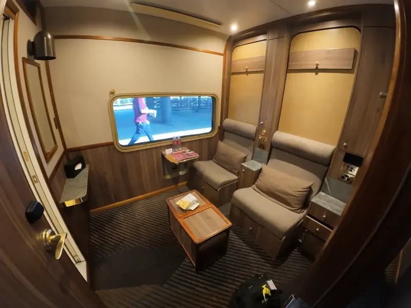 a luxurious, curved-corner room with a sophisticated design, featuring a large wooden table with the number '5' on it, a comfortable sofa with a cushion, and a large screen displaying a person walking.