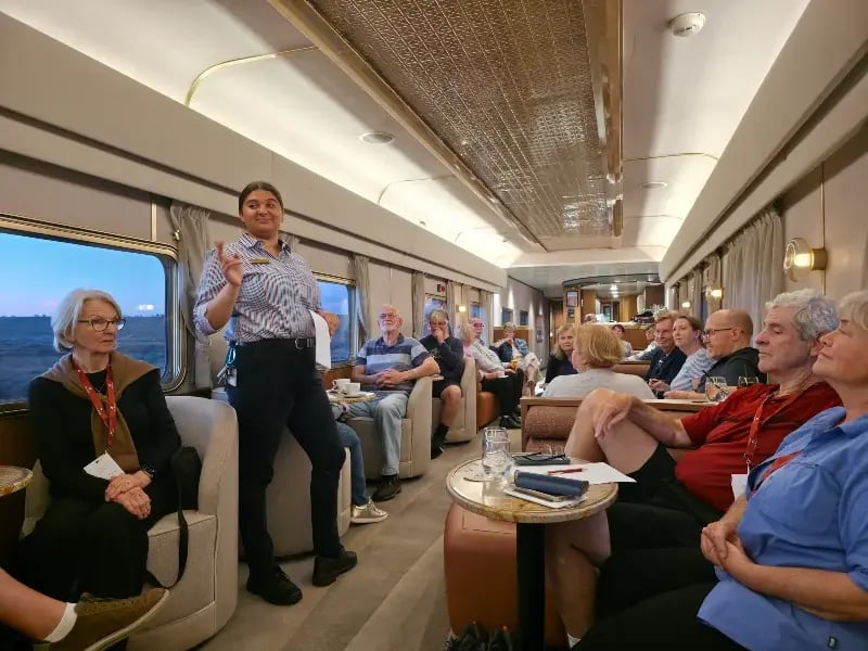 an indoor setting of a train carriage with passengers seated and standing. The carriage has a modern design with a patterned ceiling and lighting. There is a woman standing and gesturing towards the passengers, who appear to be watching her.