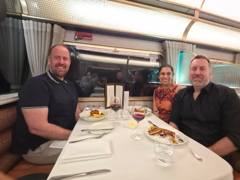 dining scene inside a vehicle, likely a train, where two men are seated across from a woman. They are surrounded by the decor and amenities of the train's dining car, which includes a table set with plates, glasses, and cutlery.