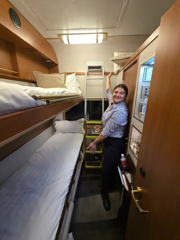 A person is standing in a small, well-lit room with multiple bunk beds, which appears to be a compact living space, possibly on a boat or ship. The person is wearing a striped shirt and dark pants, and is holding onto a pole or handle next to a door.